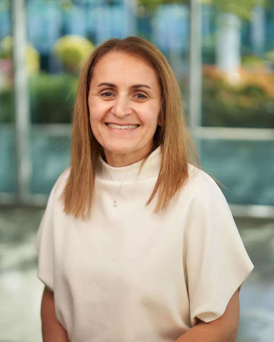 A woman wearing a white top is seated in a bright, modern office environment. The background features large windows, greenery, and blurred outdoor visuals. Her attire and surroundings suggest a professional context, with soft lighting enhancing the scene.